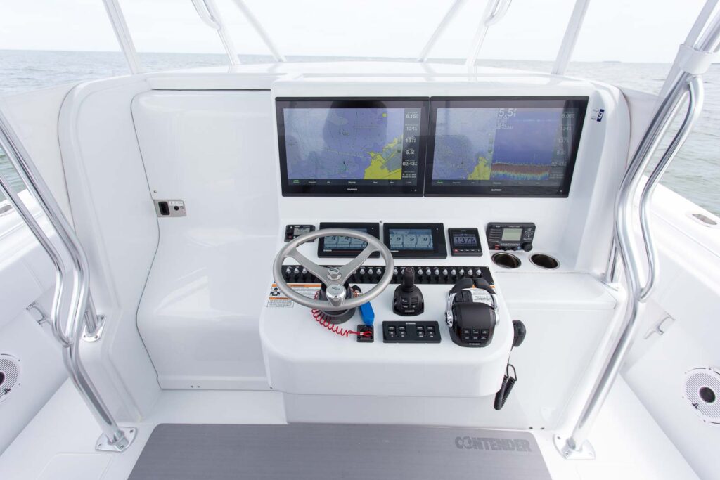 Boat cockpit with steering wheel, twin monitors displaying navigation and depth data, and various controls and instruments on a clean, white console. Ocean visible in the background.